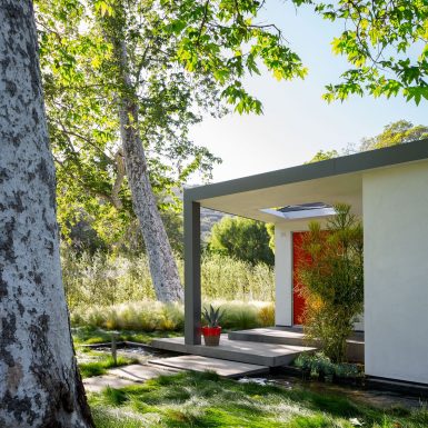 Ranch Redux - Exterior view of front porch with red front door and grassy landscape, with water feature below and sycamore trees overhead. Photograph by Trevor Tondro.